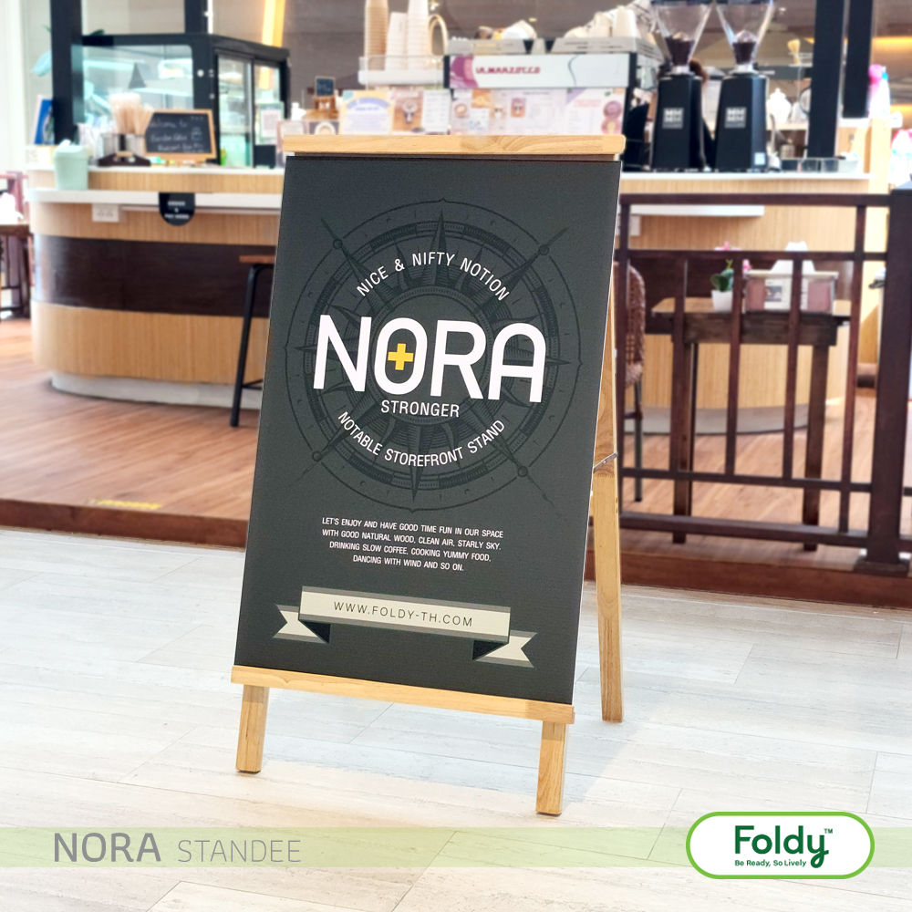 A chalkboard sign on a wooden A-frame promoting NORA—Notable storefront stand, with Foldy logo in the foreground and cafe counter in the background.