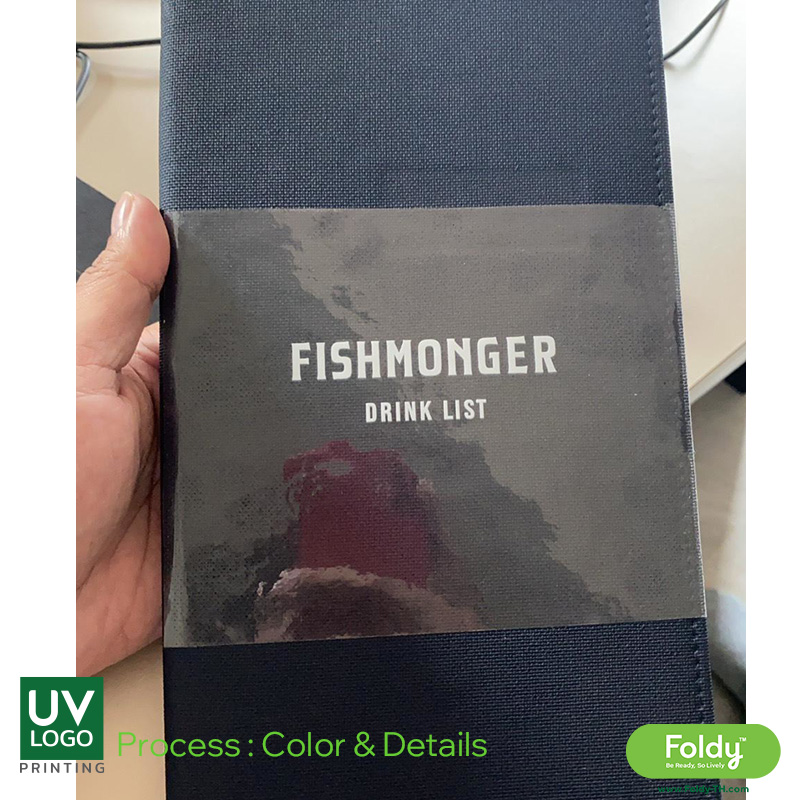 Hand holding a blue fabric booklet or case with a gray band that reads 'FISHMONGER DRINK LIST' in white letters.